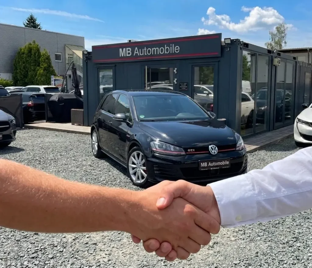 Erfolgreicher Autokauf bei MB Automobile in Weiterstadt: Käufer und Verkäufer im weißen Hemd geben sich die Hand vor einem schwarzen VW Golf GTI Performance auf dem Kiesplatz